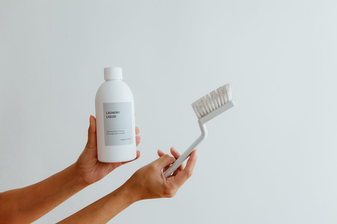 Close-up of hands holding a laundry liquid bottle and cleaning brush against a white background.