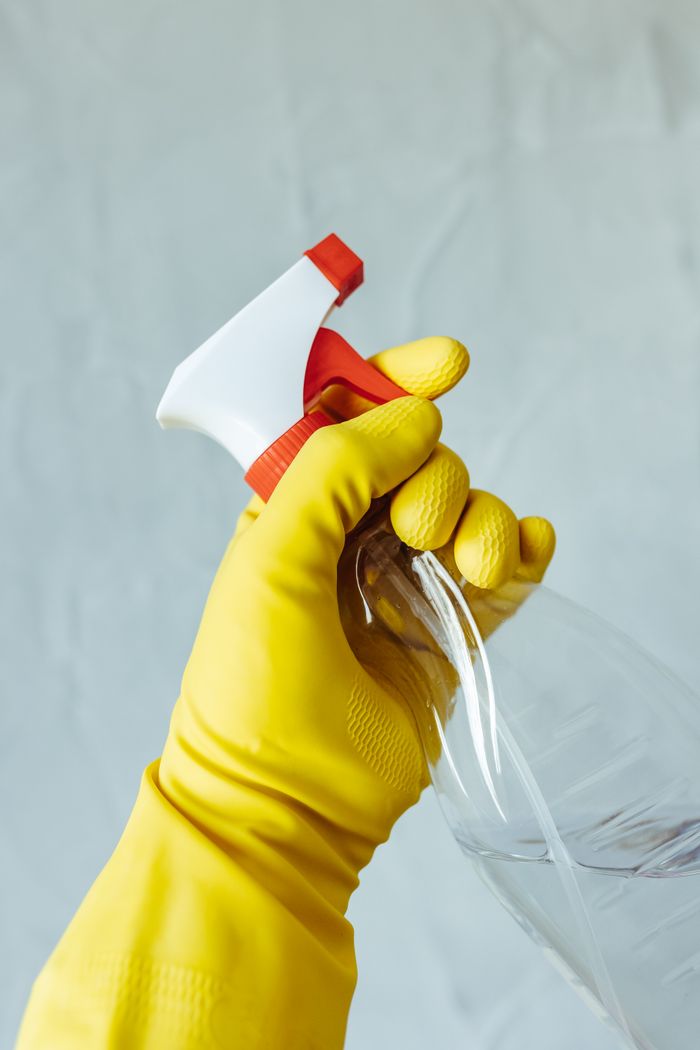why-choose-us-01 A hand wearing a yellow rubber glove holds a red and white spray bottle against a neutral background.